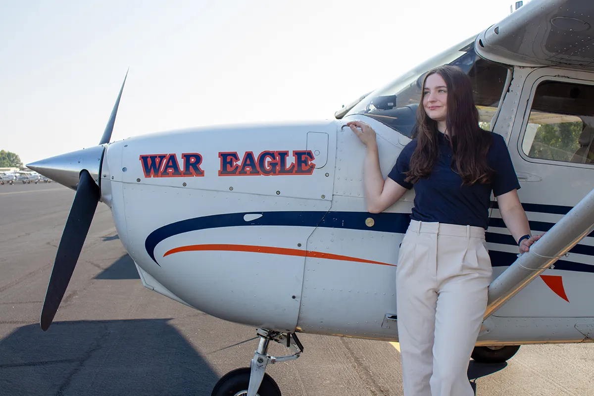 A young woman stands beside a small airplane with “War Eagle” painted on the nose, smiling while resting her hand on the aircraft wing at an airport.