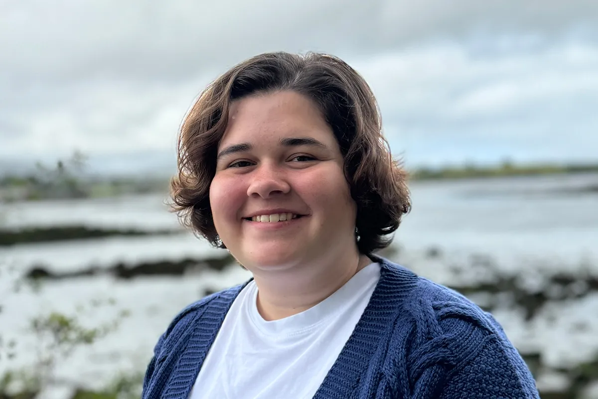 A woman with short brown hair smiles outdoors in front of a rocky shoreline, wearing a blue knit cardigan and white shirt.
