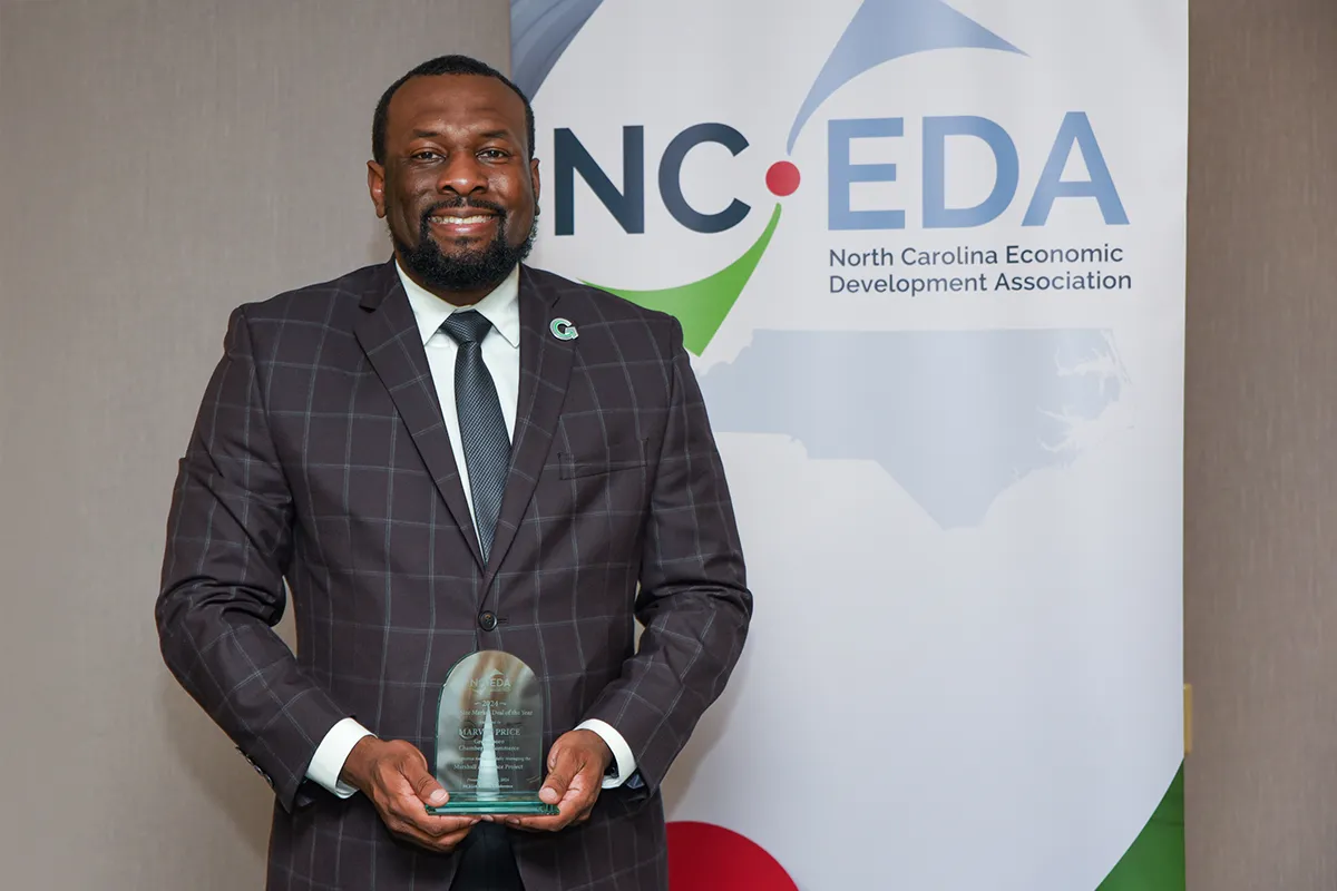A man in a dark checkered suit and tie stands in front of a banner for the North Carolina Economic Development Association, smiling and holding an award.