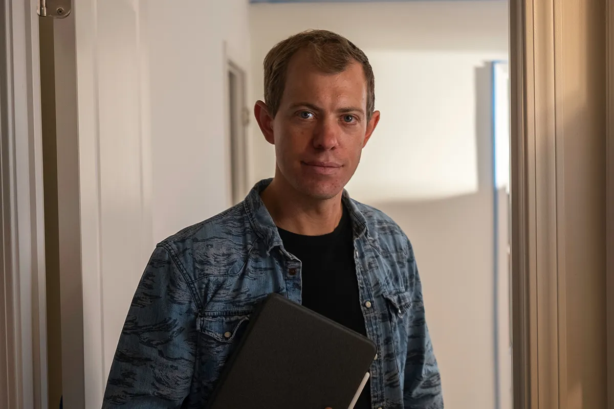 A man wearing a denim shirt holds a closed laptop or notebook while standing in a softly lit hallway, looking at the camera.