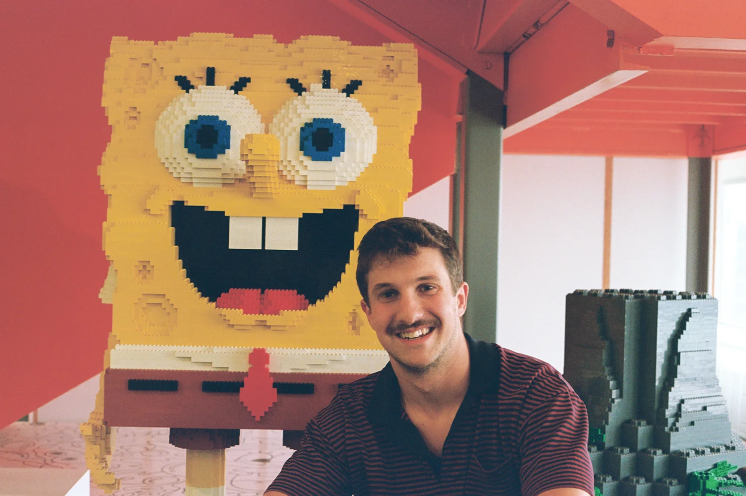 A person smiling next to a large LEGO sculpture of a cartoon character with large eyes and a wide smile.
