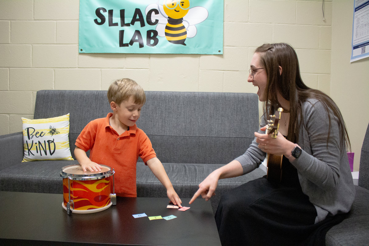 Erin Cannon playing guitar in lab singing with child client, the little boy is playing a small drum and pointing at a letter making the letter sound with music.