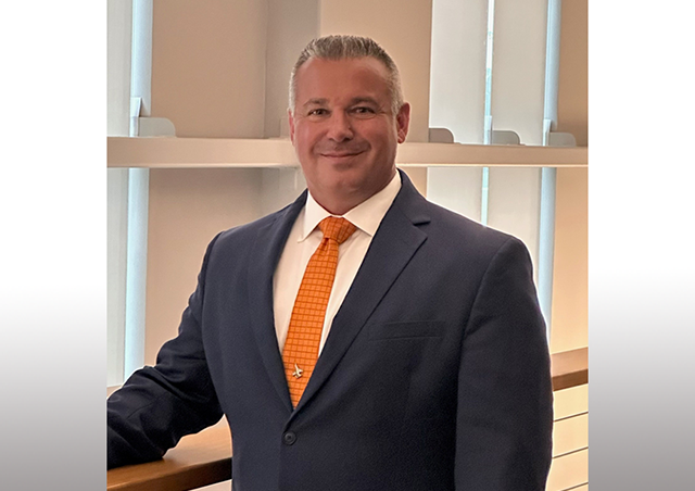 Portrait of a man wearing a navy blue suit, white shirt, and orange tie, standing indoors near a wooden railing with vertical window panels in the background.