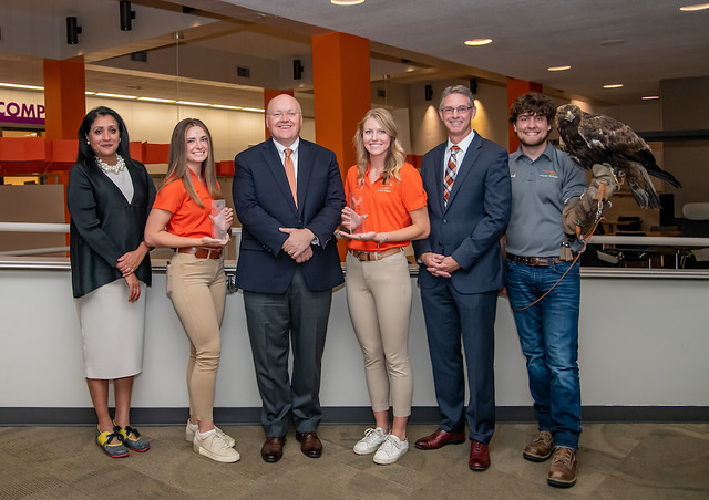 Group photo of six people standing in a modern indoor setting; two women in orange shirts hold trophies, two men in suits stand beside them, one woman in a white dress stands at the far left, and a man at the far right holds a large eagle on his gloved arm.